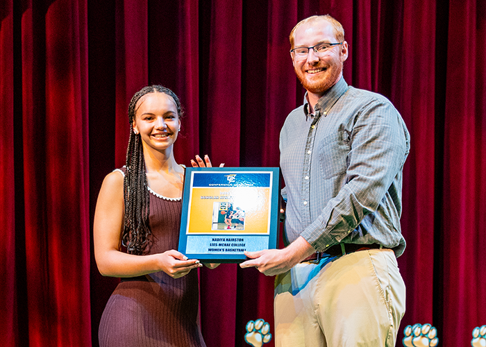Student holds award plaque with Assistant Athletic Director on stage.