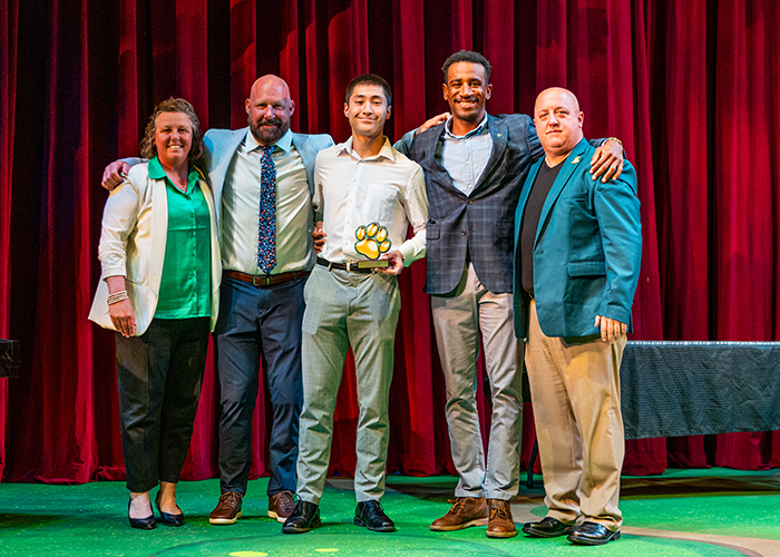 A group of students and athletics staff stand on stage while one student holds an award.