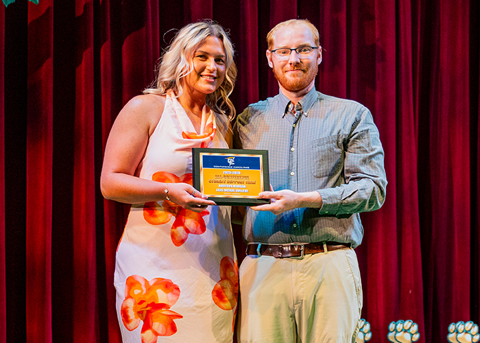 Student holds award plaque with Assistant Athletic Director on stage.