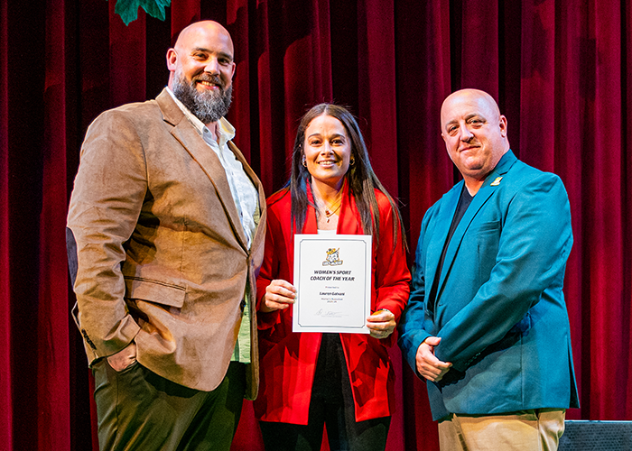 A woman stand with two men on stage while holding an award.