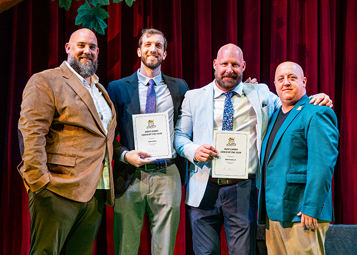 A group of men stand on stage while two of them hold awards.
