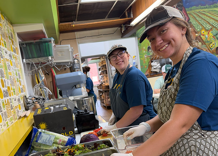 Two student volunteers smile at the camera while preparing salad greens in a kitchen.