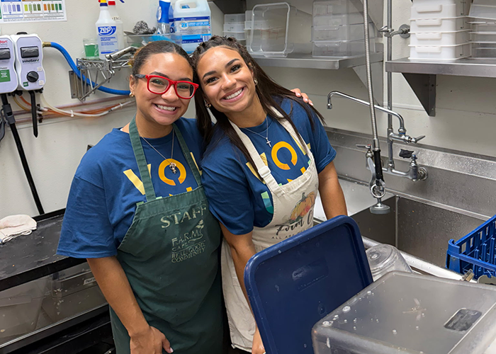 Two student volunteers smile at the camera while washing dishes in a kitchen.