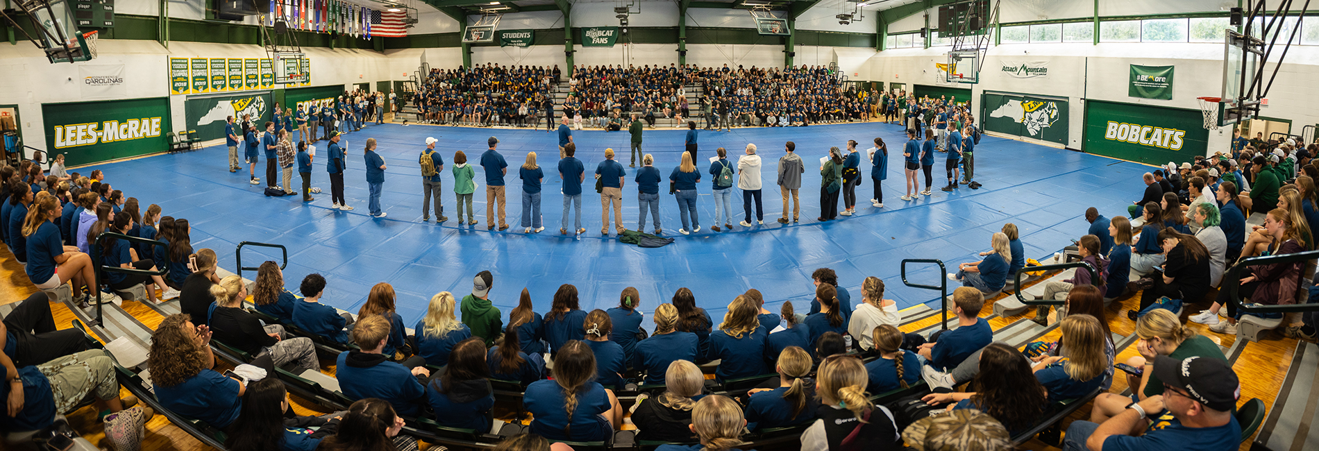 Mountain Day volunteers sit in bleachers while a group of people stand in a semi-circle on the gym floor.
