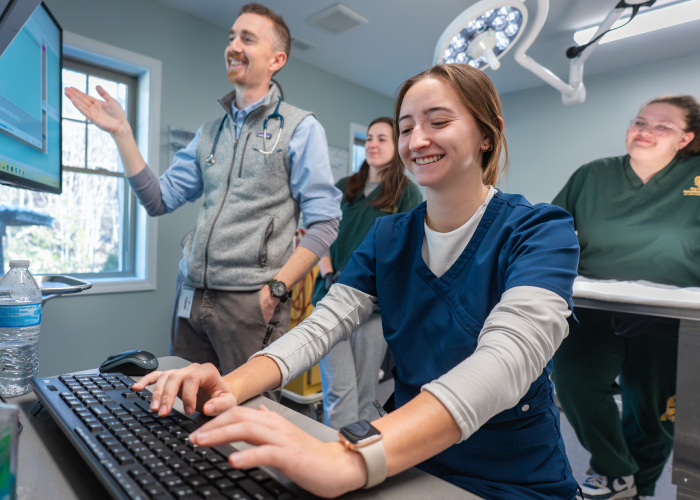 Students practice using clinical equipment while an instructor demonstrates a medical procedure.