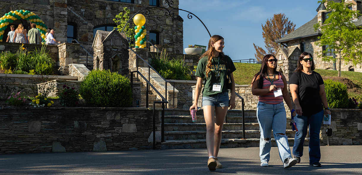 Three students walk toward the camera. North Carolina Building can be seen in the background with a green and gold balloon arch at the main entrance.