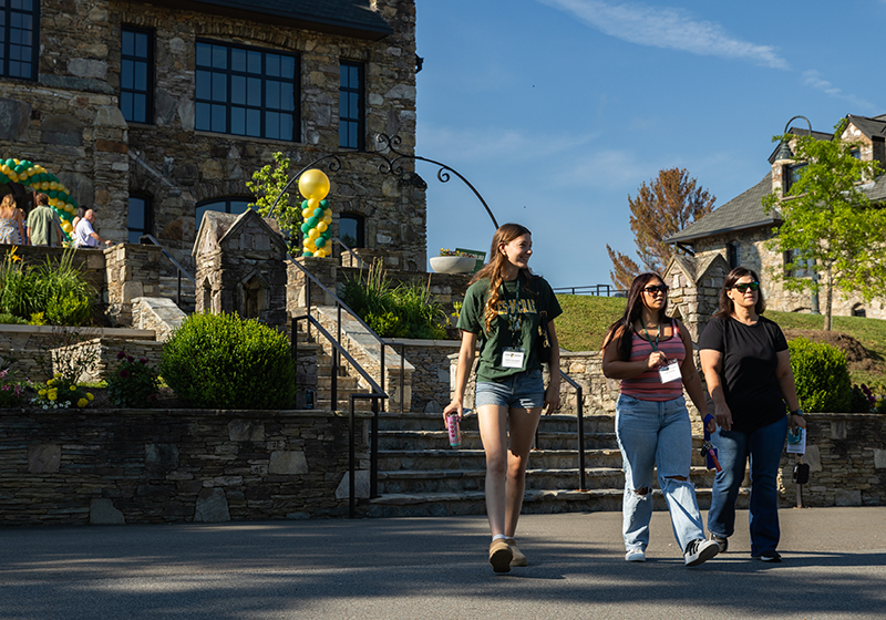 Three students walk toward the camera. North Carolina Building can be seen in the background with a green and gold balloon arch at the main entrance.