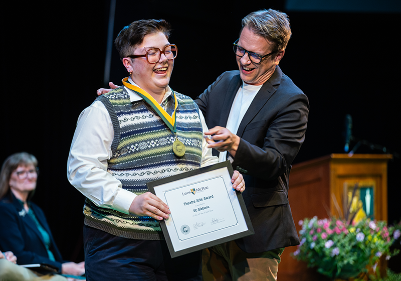 A faculty member places a medal on a smiling student who is holding a framed award certificate.