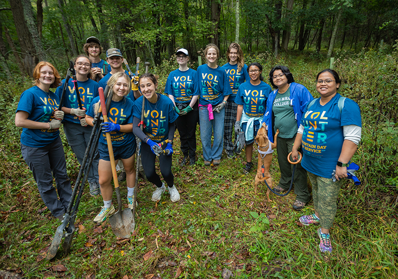 A group of volunteers wearing Mountain Day of Service shirts stand together in the woods.